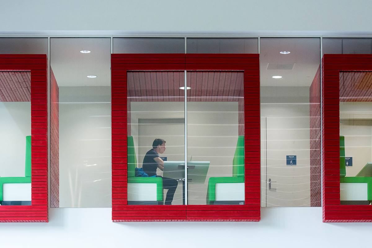 Cabine de travail vitrée avec cloisons rouges et vertes, homme assis à son ordinateur portable à l'intérieur Cabine de travail vitrée avec cloisons rouges et vertes, homme assis à son ordinateur portable à l'intérieur