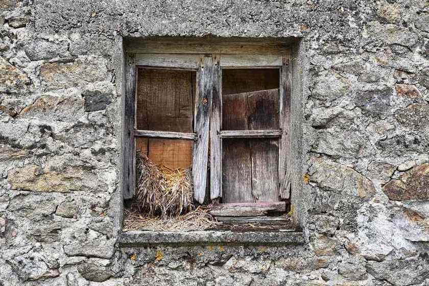 Fenêtre en bois décrépit enchâssée dans un mur en pierre grise et fissurée, avec du foin visible à l'intérieur