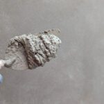 Une main gantée tient une spatule chargée d'enduit sur un mur gris